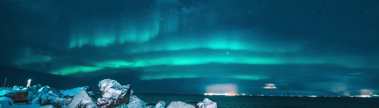 The Northern Lights illuminate the sky above a bay, with snow-covered rocks in the foreground