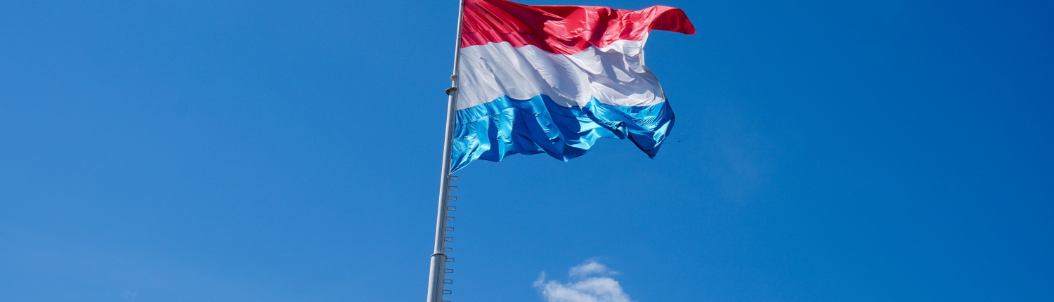 The red-, white- and blue-striped Luxembourg flag flying in front of a blue sky